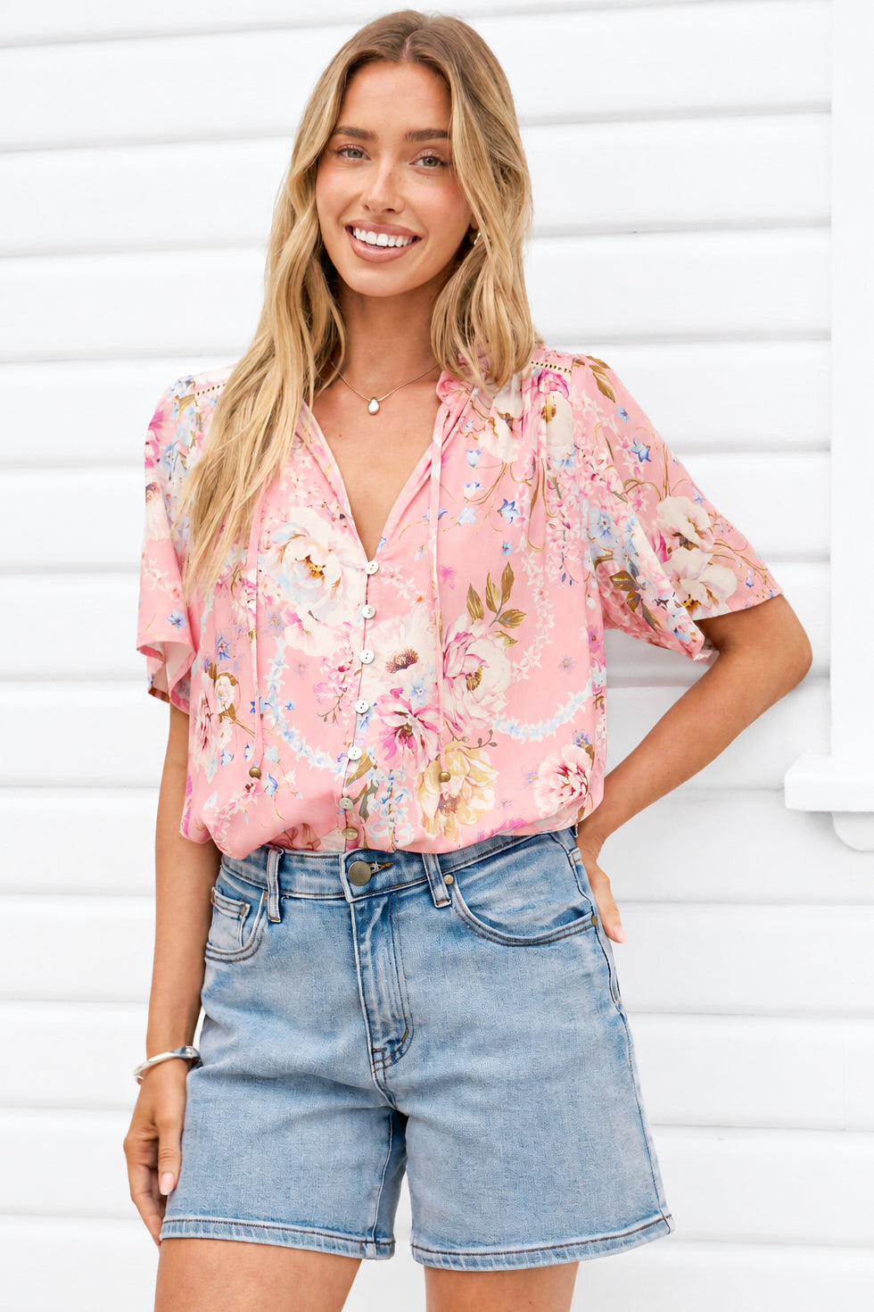 A woman wearing a light pink floral short-sleeve blouse and light-wash denim shorts, standing against a white slatted wall.