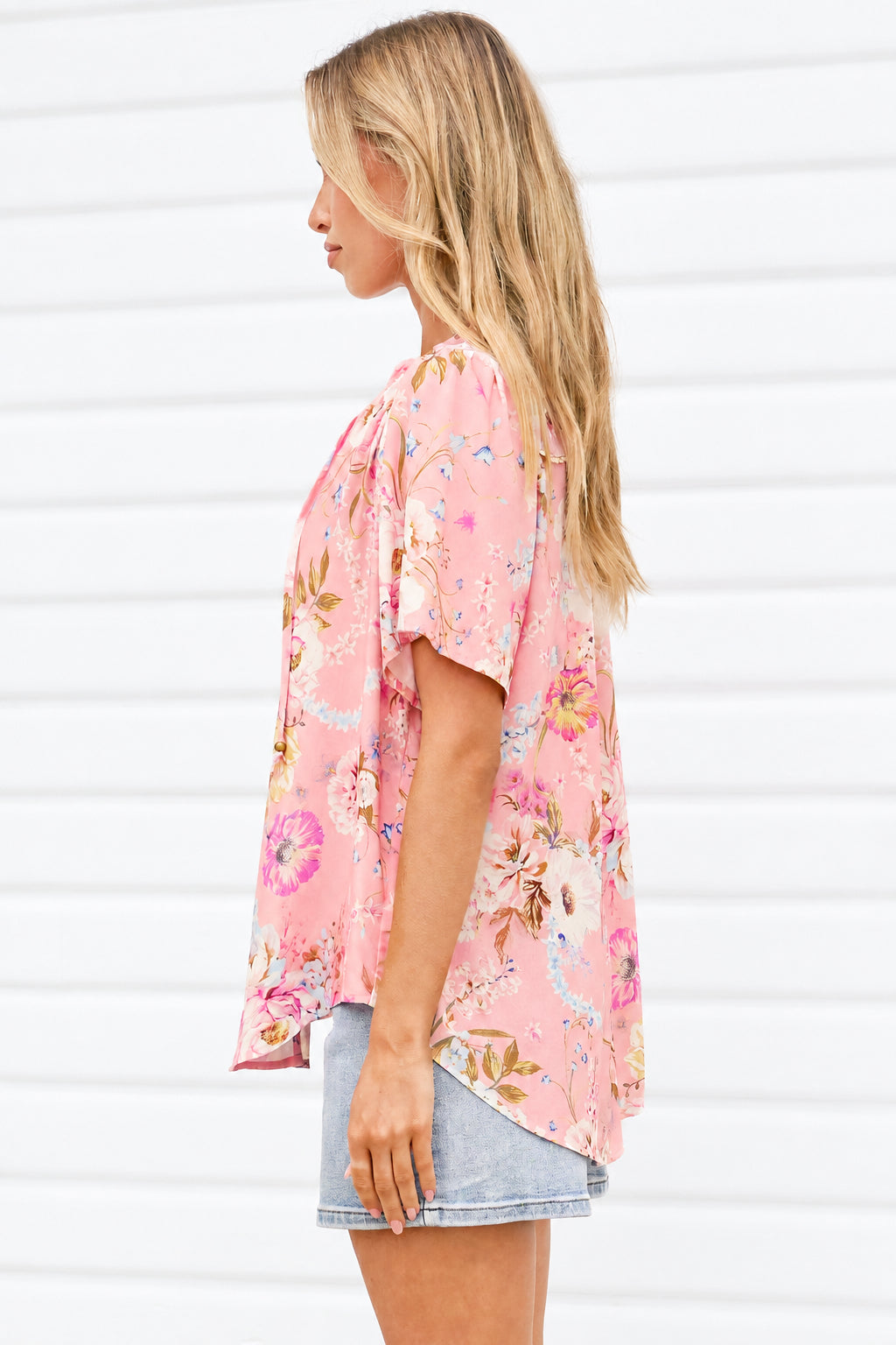 A woman shown from the side profile wearing a pink short-sleeve floral top and denim shorts, standing against a white paneled wall.