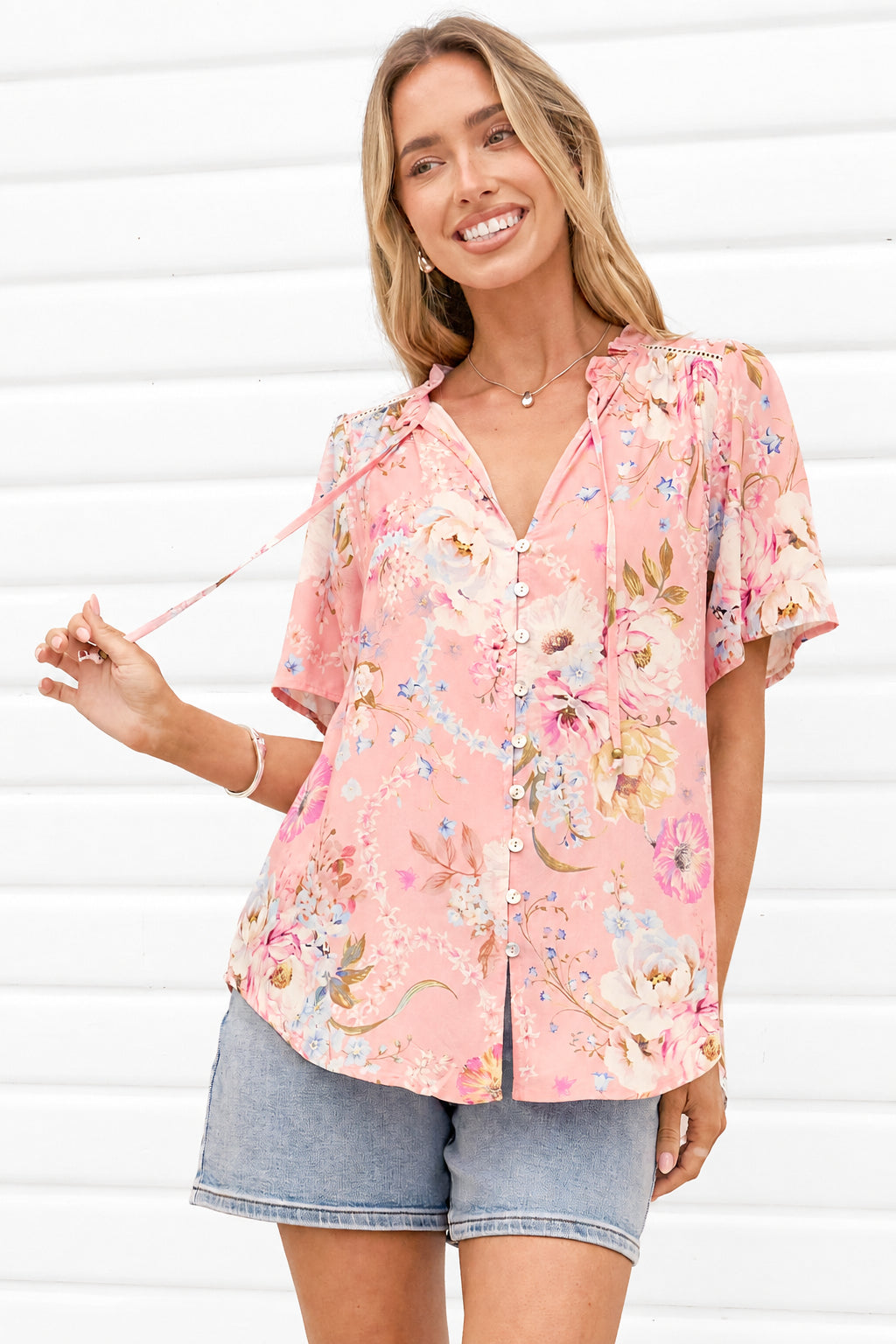 A woman wearing a pink short-sleeve button-up blouse with a colorful floral print and denim shorts, standing against a white paneled wall.