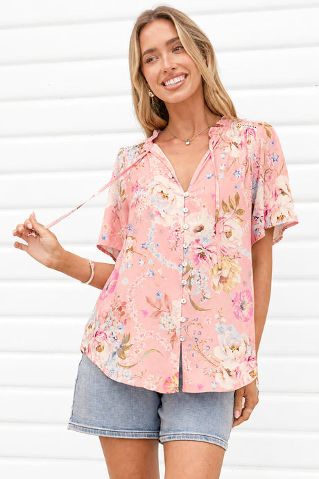 A woman wearing a pink short-sleeve button-up blouse with a colorful floral print and denim shorts, standing against a white paneled wall.