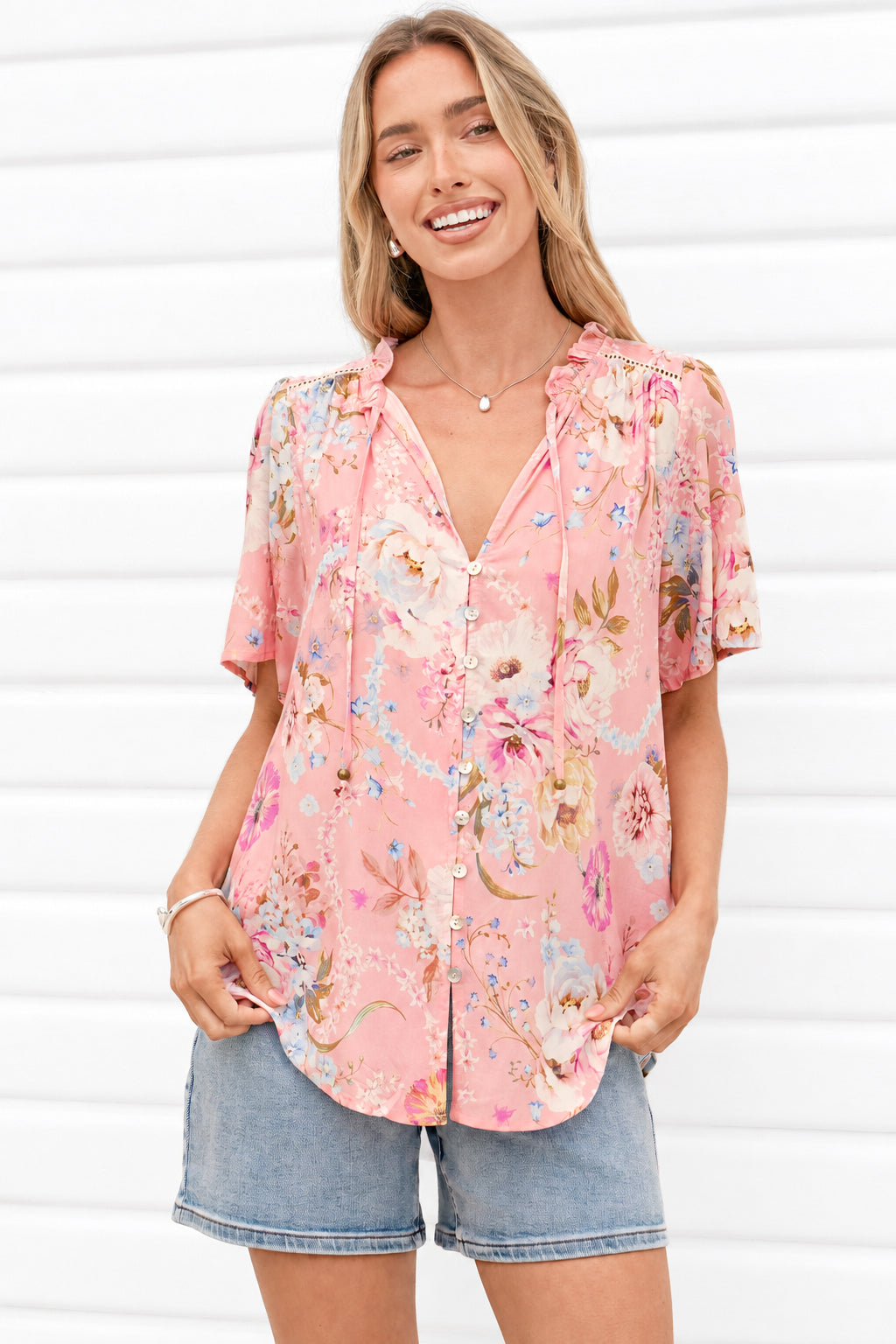 A woman wearing a pink floral short-sleeve button-up blouse and denim shorts, standing against a white paneled wall.