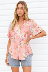 A woman wearing a pink floral short-sleeve button-up top and denim shorts, standing against a white slatted wall.