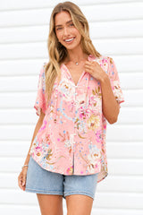 A woman wearing a pink short-sleeve button-up blouse with a colorful floral and paisley print, paired with denim shorts, standing against a white paneled wall.