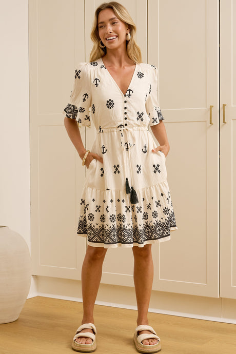 A woman in a relaxed beige printed mini dress featuring anchor and geometric motifs, a button-front neckline, and tasselled waist tie, posed in front of a warm beige wardrobe.