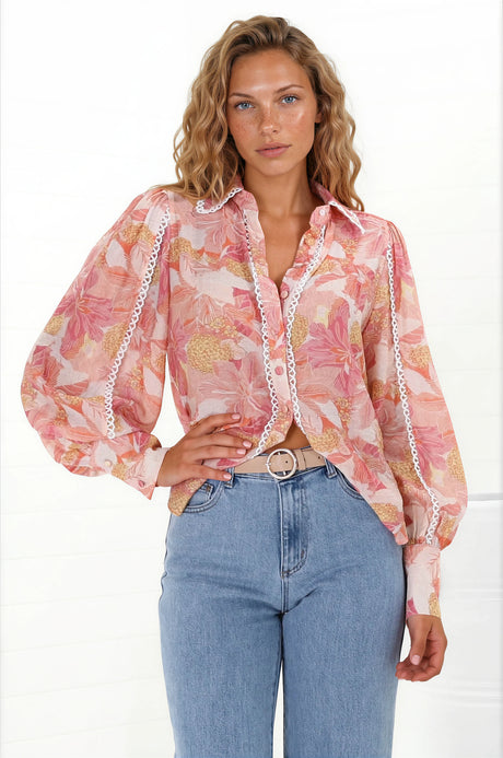 A woman wearing a pink and orange floral button-down blouse with contrast trim lace detailing, with long sleeves and a scoop hemline, paired with blue jeans.