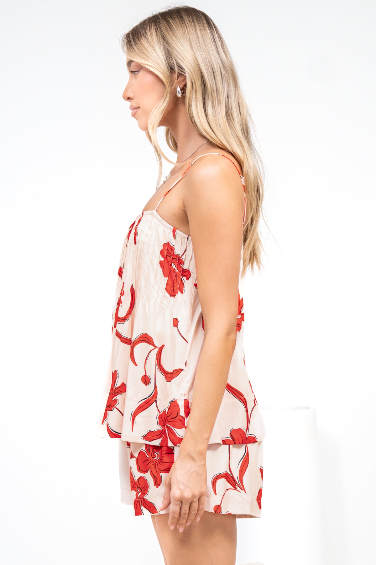 Side view of a woman wearing the Soleia Playsuit in Arli Print, a spaghetti strap cami overlay playsuit in a cream and red bold floral print, against a white wall