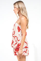 Side view of a woman wearing the Soleia Playsuit in Arli Print, a spaghetti strap cami overlay playsuit in a cream and red bold floral print, against a white wall