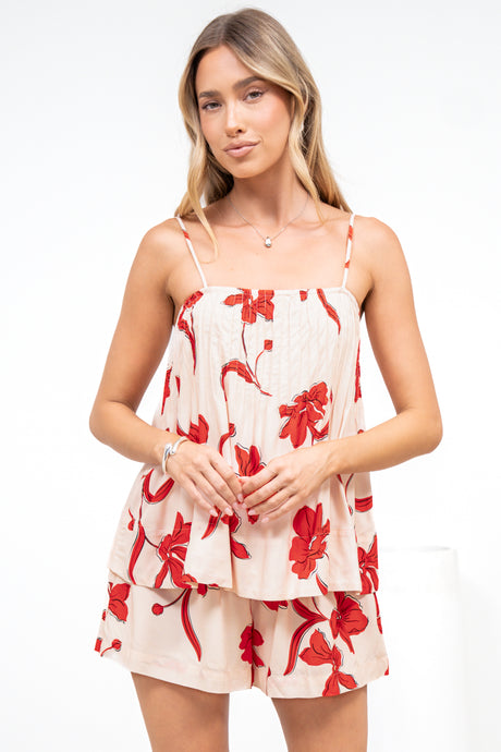 A woman wearing the Soleia Playsuit in Arli Print, hands clasped in front of the pin tuck cami overlay playsuit in a cream and red bold floral print, against a white wall