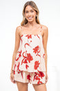 A smiling woman wearing the Soleia Playsuit in Arli Print, a spaghetti strap playsuit with a pin tuck cami overlay in a cream and red bold floral print, against a white wall