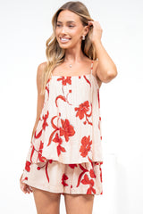 A smiling woman touching her hair, wearing the Soleia Playsuit in Arli Print, a spaghetti strap cami overlay playsuit in a cream and red bold floral print, against a white wall