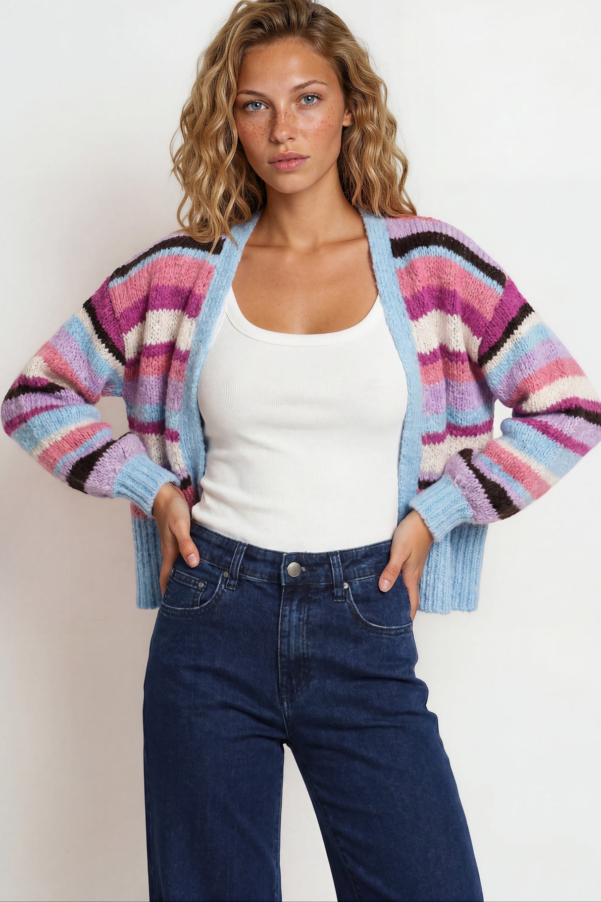 Woman wearing a colorful striped cardigan over a white tank top and blue jeans against a plain background