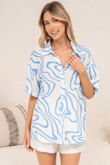 A woman with one hand at her collar wearing the Sutton Shirt in Spensley Print, an oversized short-sleeve button-down in a blue and white wavy swirl print, styled loose over white shorts in front of a cream sofa