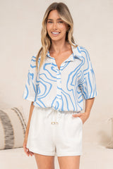 A smiling woman with her hand in her pocket wearing the Sutton Shirt in Spensley Print, a collared short-sleeve button-down in a blue and white wavy swirl print, tucked into white drawstring shorts in front of a cream sofa