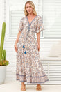 A woman modeling a bohemian-style maxi dress with a paisley pattern in beige, blue, and brown, featuring flutter sleeves and blue tassel ties, set against a bright white porch background.