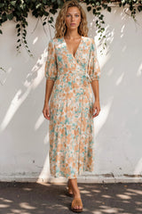 Woman wearing a floral dress standing against a white wall with greenery.