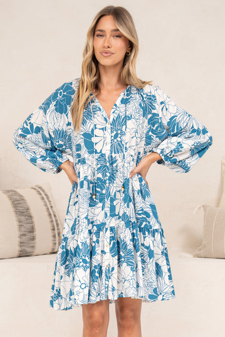 A woman with her hands on her waist wearing the Tia Mini Dress in Lumina Print, a white and blue tropical leaf print V-neck mini dress with long balloon sleeves and a tiered hem, in front of a cream sofa with neutral cushions