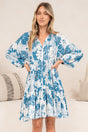 A woman with her hands on her waist wearing the Tia Mini Dress in Lumina Print, a white and blue tropical leaf print V-neck mini dress with long balloon sleeves and a tiered hem, in front of a cream sofa with neutral cushions