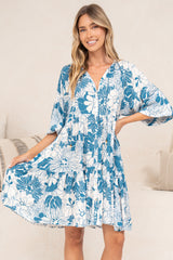 A smiling woman holding out the tiered skirt of the Tia Mini Dress in Lumina Print, a white mini dress with a blue tropical leaf print, V-neckline and flared sleeves, standing in front of a cream sofa with neutral cushions