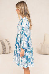 Side view of a woman wearing the Tia Mini Dress in Lumina Print, showing the relaxed fit, long sleeves and tiered mini hem of the blue and white tropical leaf print dress, in front of a cream sofa with neutral cushions
