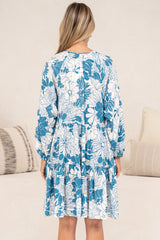 Back view of a woman wearing the Tia Mini Dress in Lumina Print, showing the loose back, long balloon sleeves and tiered mini hem in a blue and white tropical leaf print, in front of a cream sofa with neutral cushions