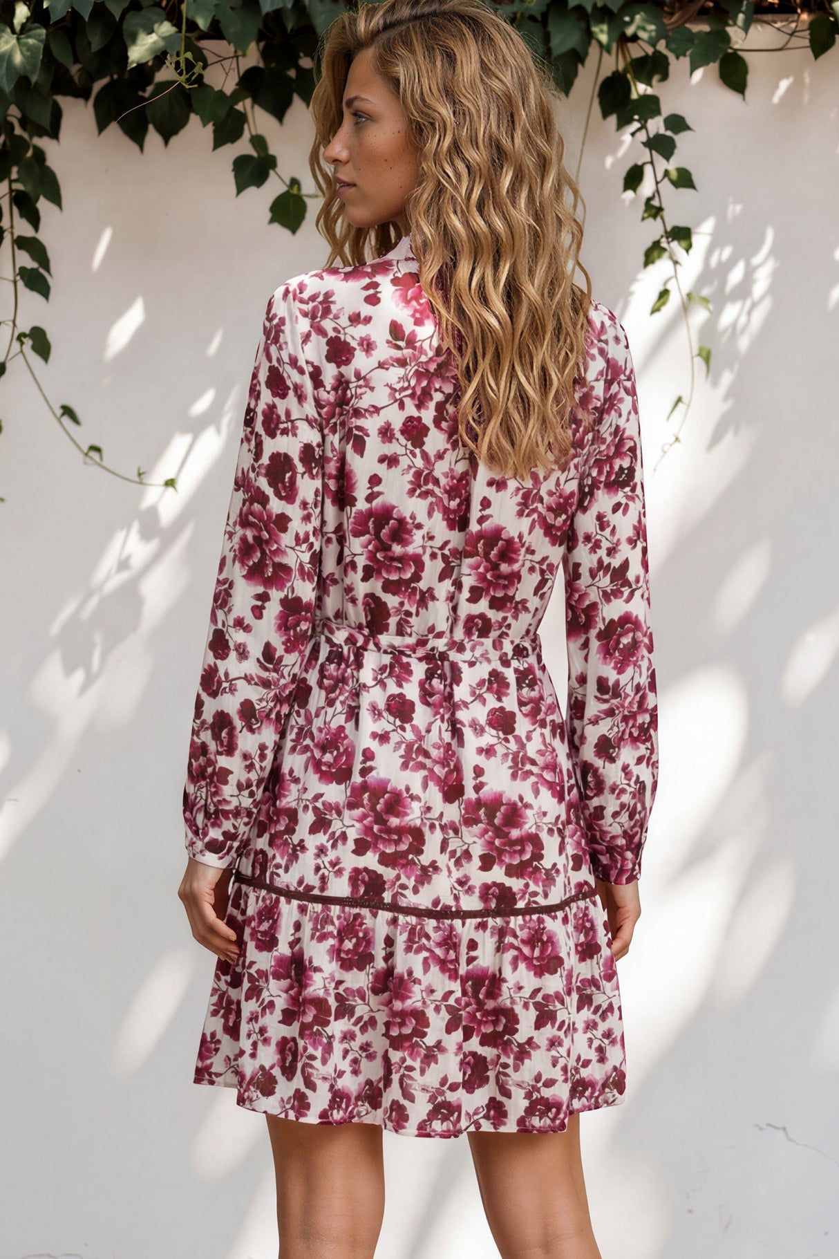 Woman wearing a floral dress standing against a white wall with greenery.