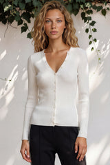 Woman wearing a white cardigan against a white wall with greenery.