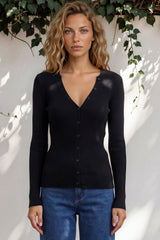 Woman wearing a black cardigan and blue jeans against a white wall with greenery.
