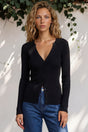Woman wearing a black cardigan and blue jeans against a white wall with greenery.
