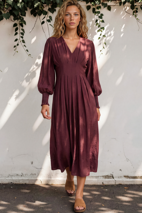 Woman in a burgundy dress standing against a white wall with greenery.