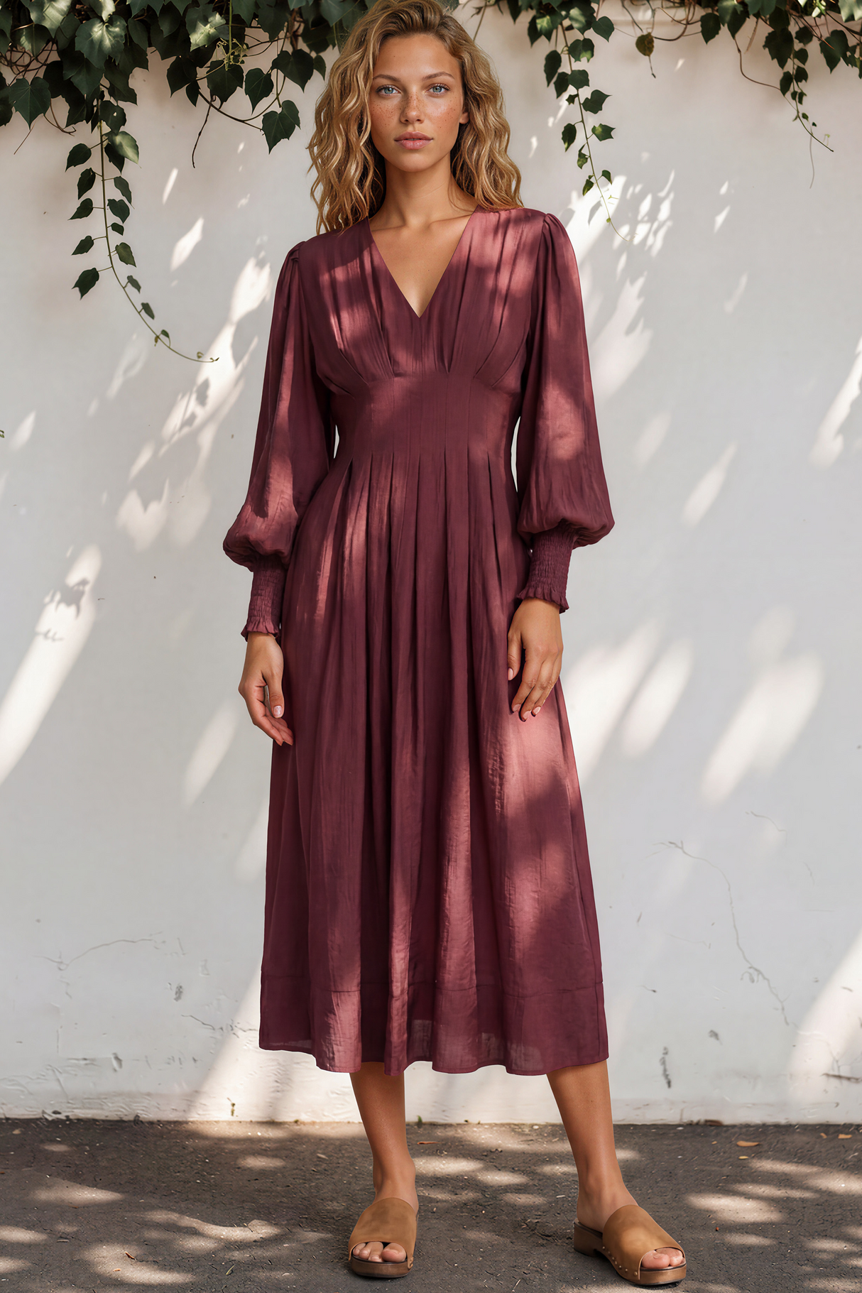 Woman wearing a burgundy dress standing against a white wall with greenery.
