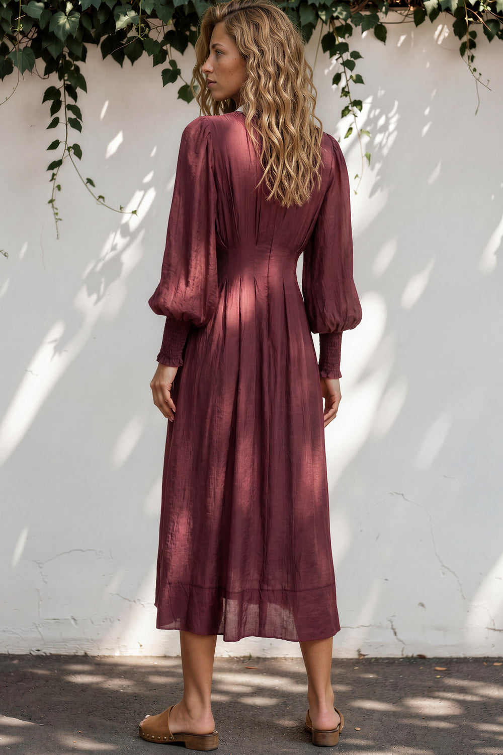Woman wearing a burgundy dress standing against a white wall with greenery.