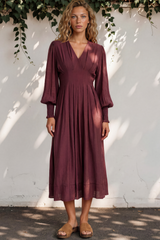 Woman wearing a burgundy dress standing against a white wall with greenery.