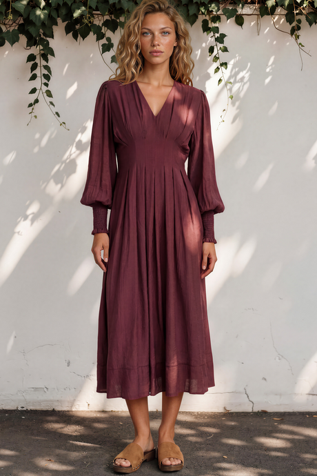 Woman wearing a burgundy dress standing against a white wall with greenery.