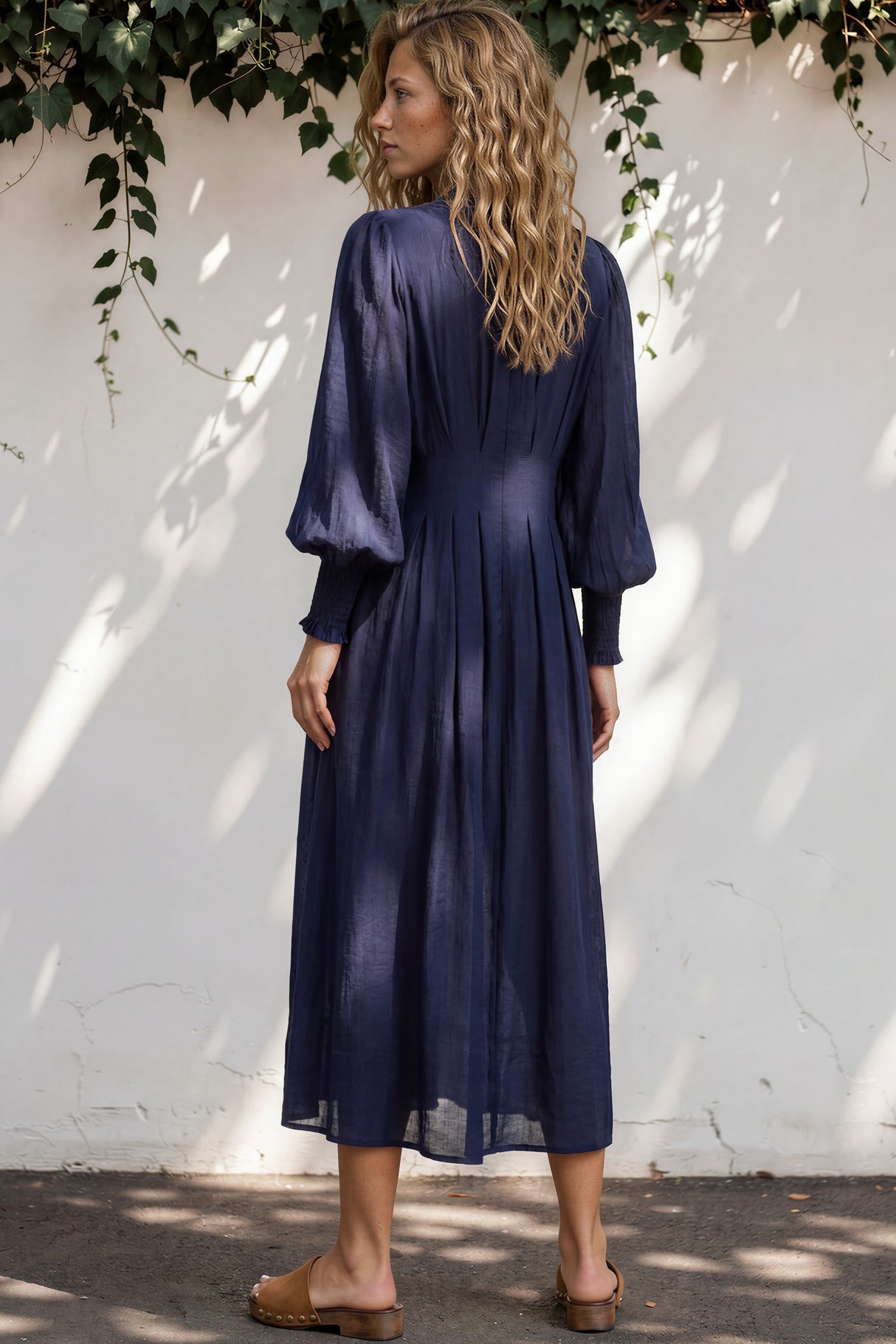 Woman wearing a long navy dress standing against a white wall with greenery.