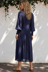 Woman wearing a long navy dress standing against a white wall with greenery.