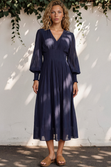 Woman wearing a navy blue dress standing against a white wall with greenery.