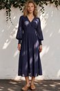 Woman wearing a navy blue dress standing against a white wall with greenery.