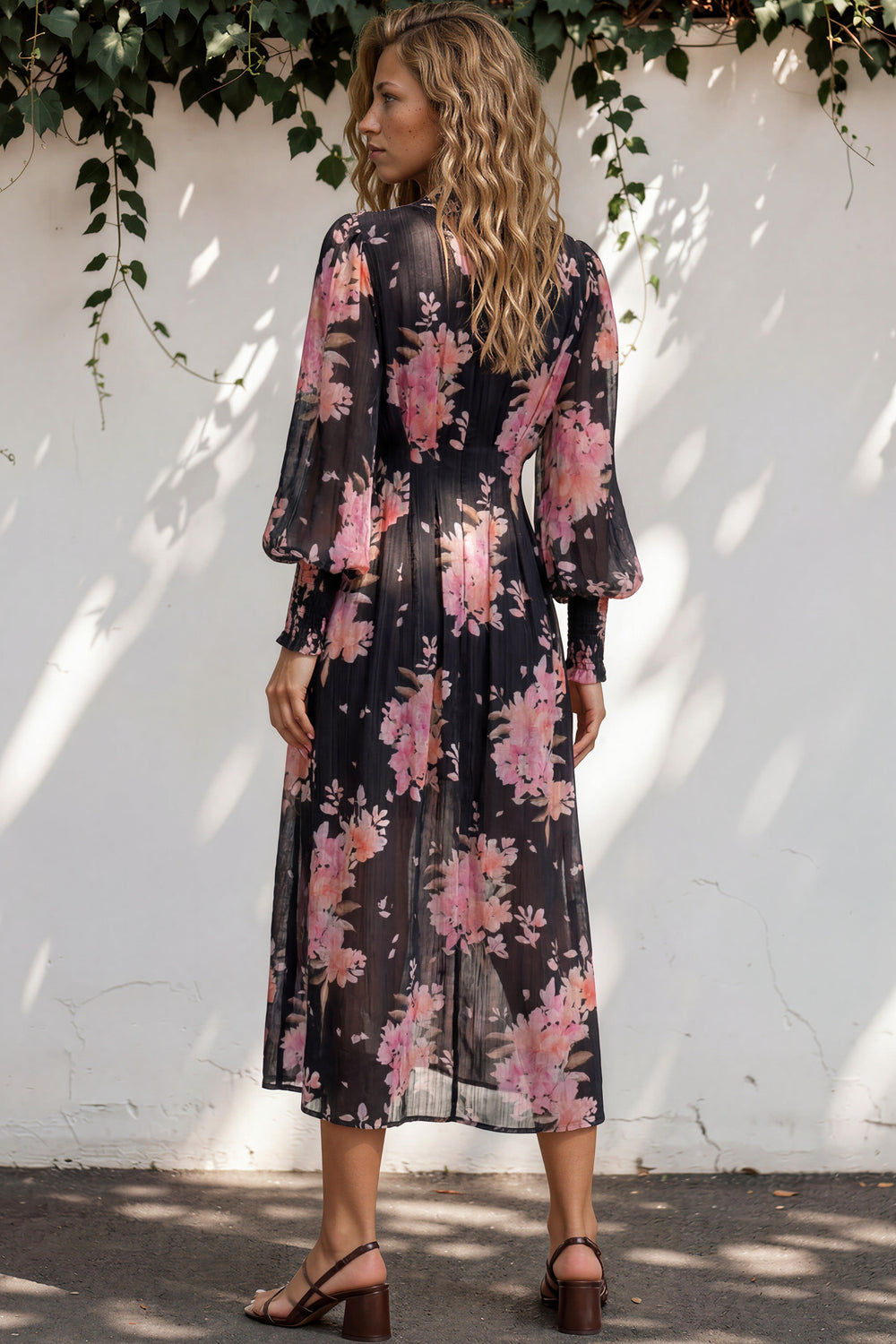 Woman wearing a floral dress standing against a white wall with greenery.