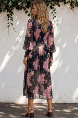 Woman wearing a floral dress standing against a white wall with greenery.