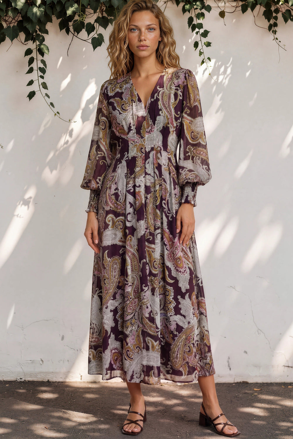 Woman wearing a patterned dress standing against a white wall with greenery.