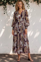 Woman wearing a patterned dress standing against a white wall with greenery.