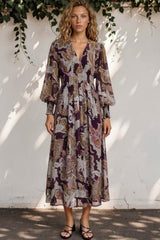 Woman wearing a patterned dress standing against a white wall with greenery.