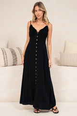A woman wearing a black maxi dress with a sweetheart neckline and shoulder ties, standing in front of a cream wall with a matching couch behind her.