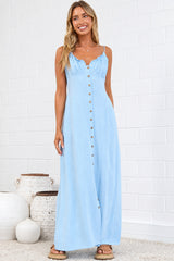 A woman modeling a light blue sleeveless maxi dress with a button-down front, standing in a bright room with white brick walls, a beige rug, and decorative ceramic vases.