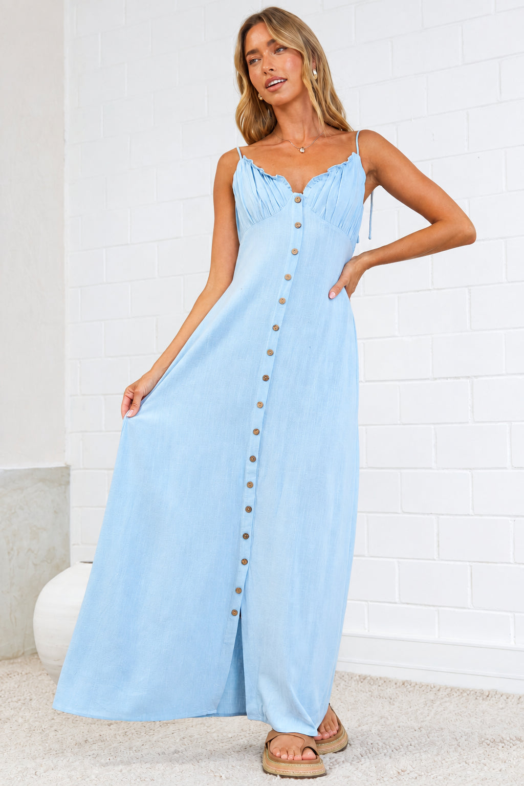 A woman modeling a light blue maxi dress with spaghetti straps and a button-down front, standing on a beige rug against a white brick wall. 