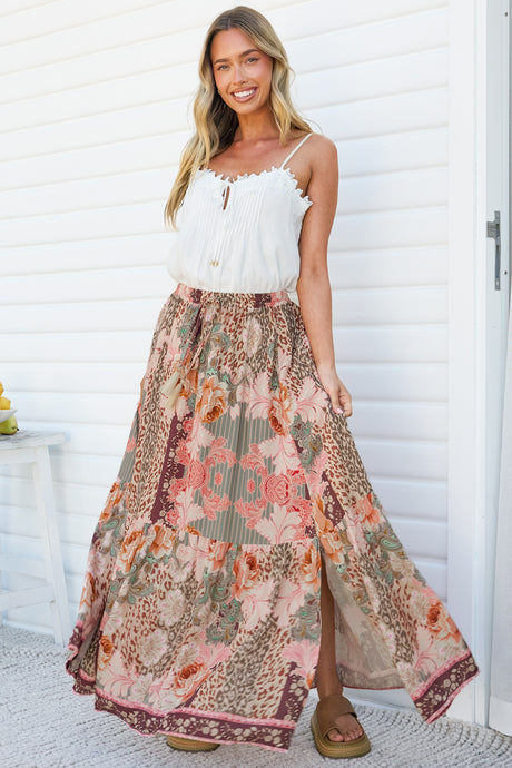 A smiling woman wearing the Zia Maxi Skirt in Frannie Print, a high-waisted maxi skirt with a tassel waist tie and front split in a pink, sage and burgundy floral patchwork print, paired with a white cami against a white wall
