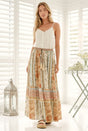 A woman modeling a white sleeveless top and a long, wide-leg maxi skirt with a multicolor floral and paisley print in beige, orange, and blue tones, standing in front of a white shuttered window.