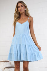 A woman modeling a light blue tiered spaghetti-strap dress in a bright, modern studio with a white brick wall and neutral decor.