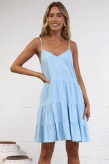 A woman modeling a light blue sleeveless mini dress with a tiered skirt, standing on a textured rug in a bright, modern room with a white brick wall. 