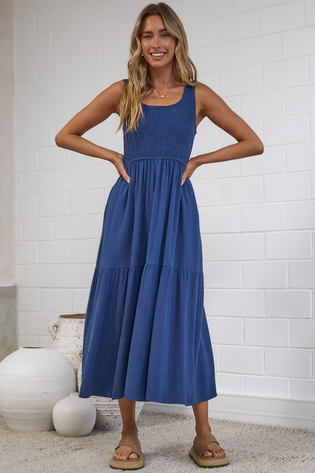 A woman modeling a sleeveless, tiered blue maxi dress in a bright, modern studio with white brick walls and neutral decor.